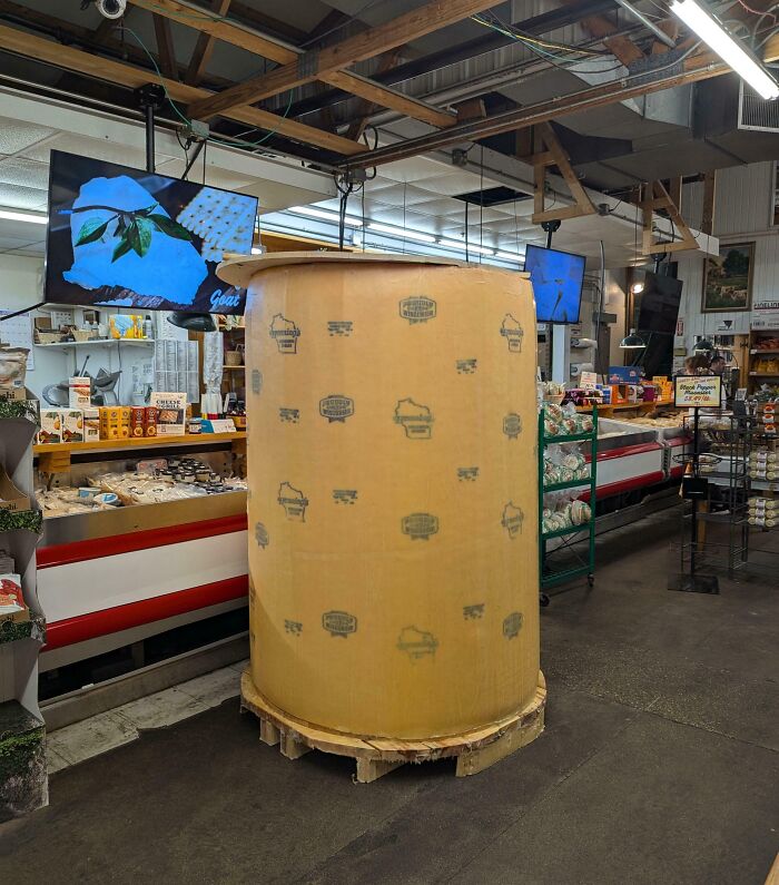 Giant cheese wheel displayed in a grocery store aisle, surrounded by various food items and digital screens overhead.