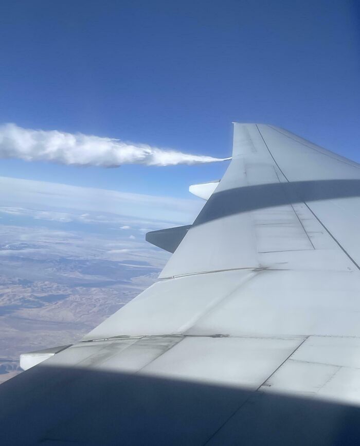 View of an airplane wing with a shadow and clouds, captured from a window, showcasing fascinating aerial scenery.