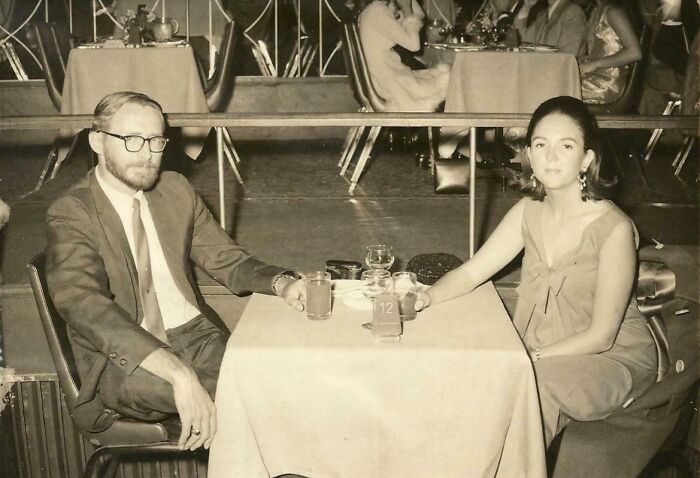 A couple at a table in 60s fashion looks, with the man in a suit and tie, and the woman in a sleeveless dress.