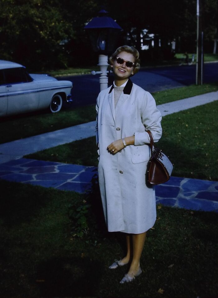 Woman in vintage 60s fashion wearing a white coat, sunglasses, and holding a purse, standing outdoors near a classic car.