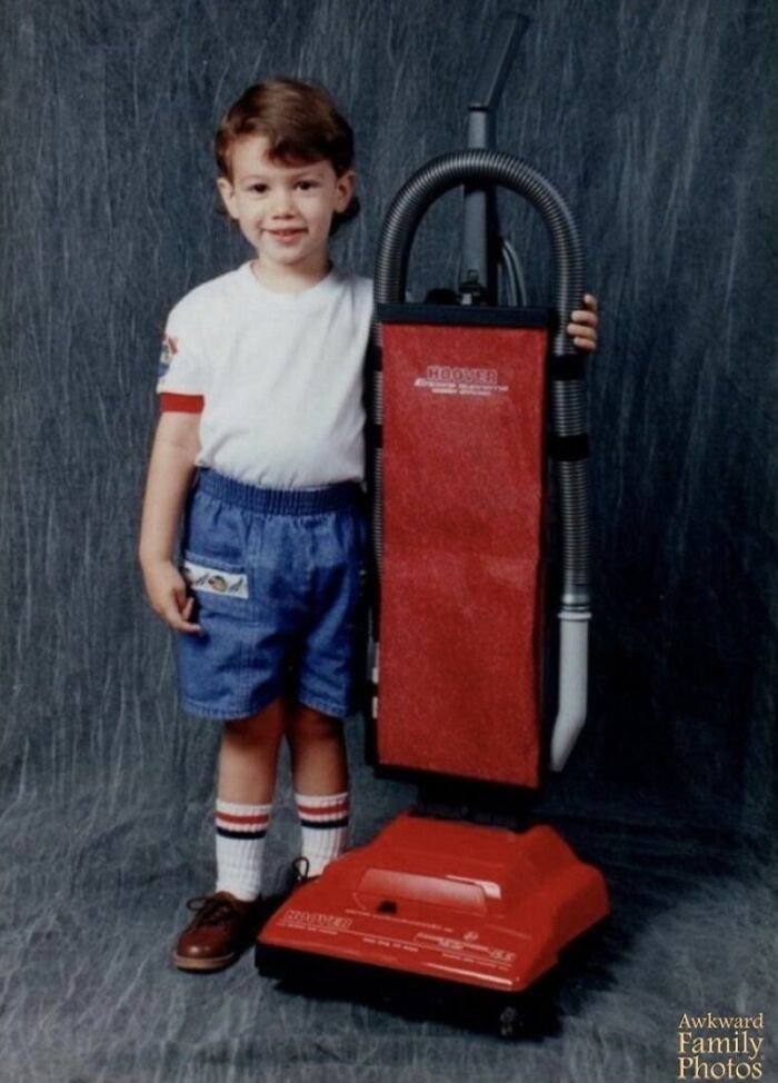 Awkward family photo of a child smiling next to a red vacuum cleaner, wearing a white shirt and blue shorts.