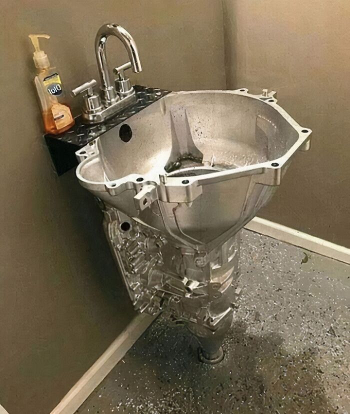 Unhinged pic of a sink made from a car engine part, with a faucet and soap dispenser on a speckled floor.