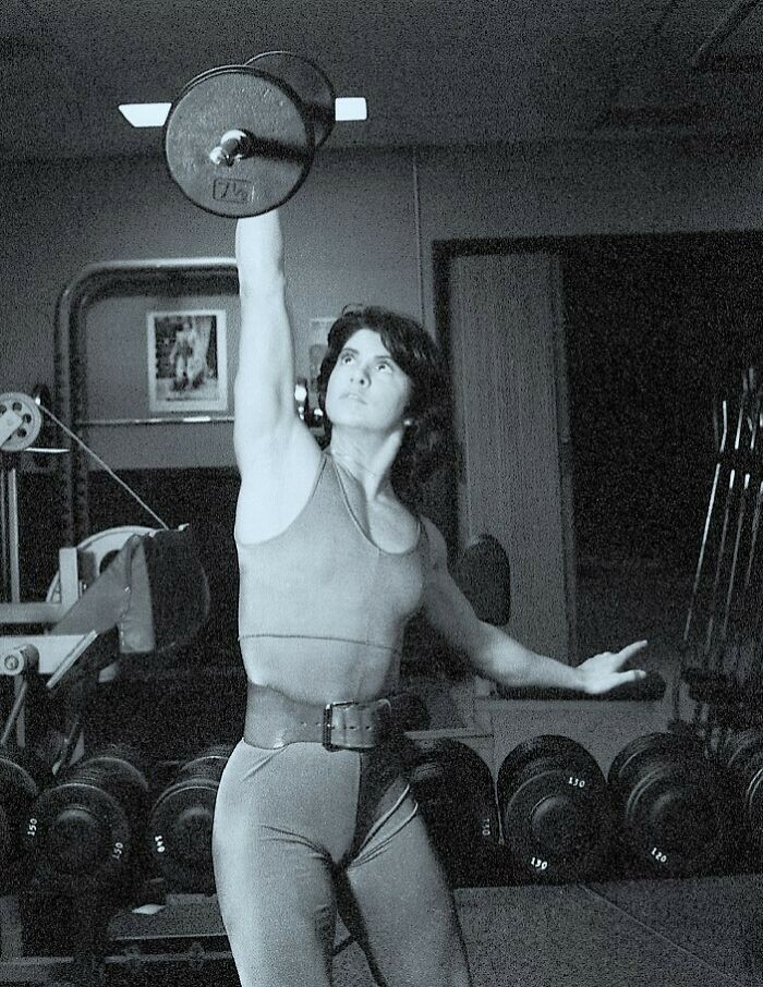 1980s gym-goer lifting a dumbbell with one hand in a fitness studio.