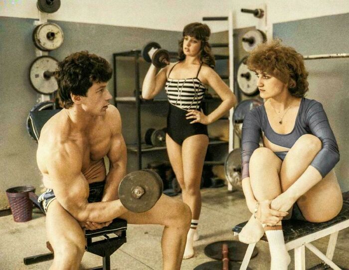 Three people in 80s gym attire with dumbbells and workout gear in a fitness studio setting.