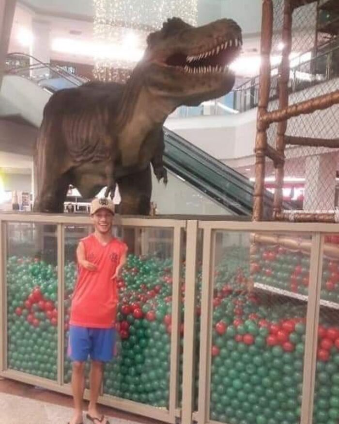 A boy in a mall poses with a dinosaur statue above a ball pit, creating a cursed image effect.