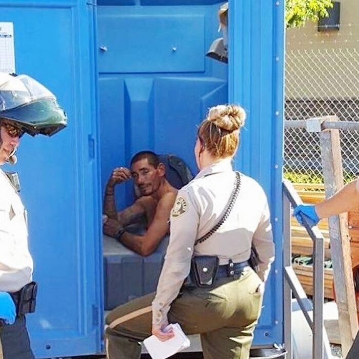 Two officers interact with a man inside a blue portable restroom in a public setting. Cursed images theme.