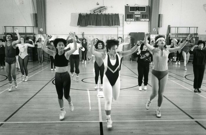 People in an 80s gym fitness class, wearing retro workout attire, exercising in a gymnasium.