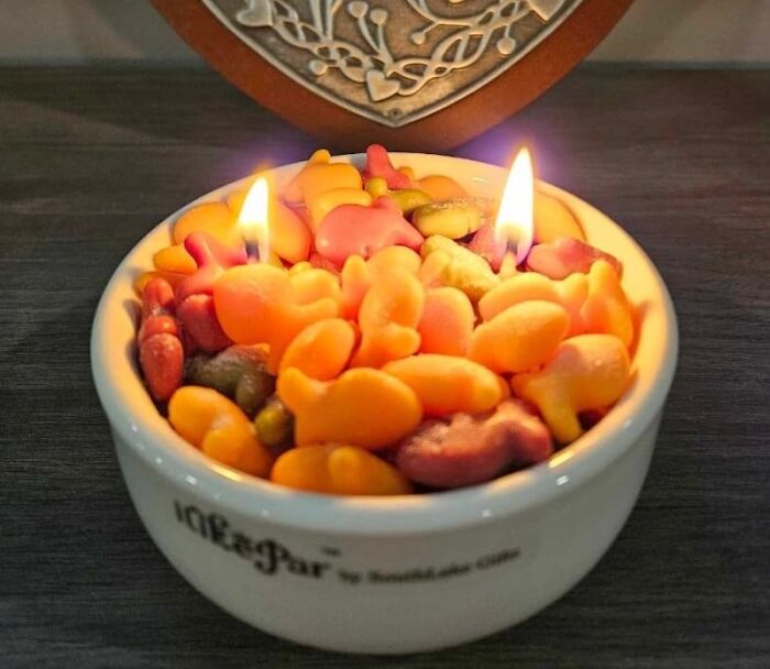 Colorful fish-shaped candles in a white bowl, burning, with a decorative background.