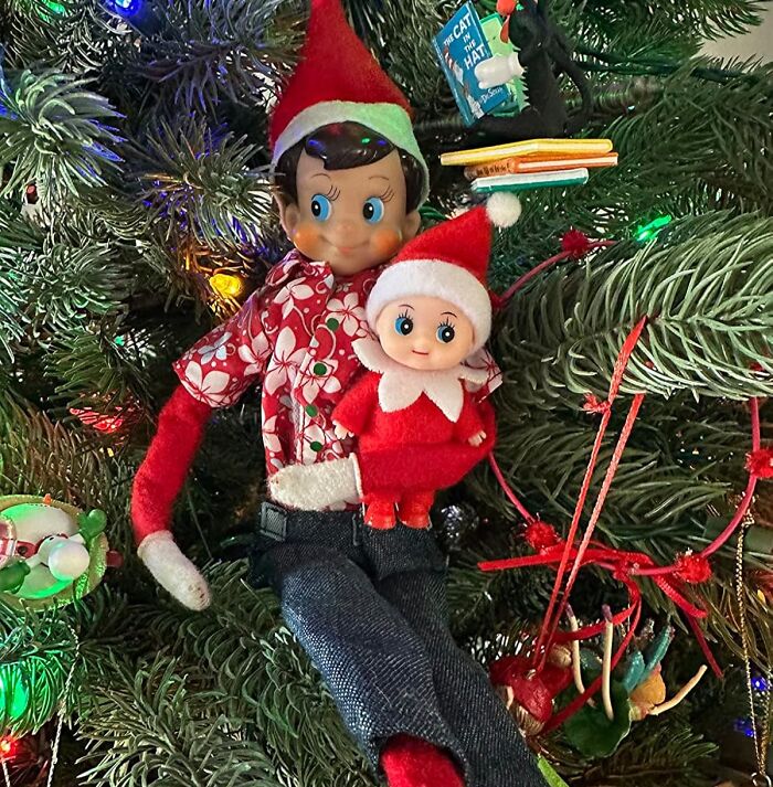 Elf dolls in festive outfits sit on a decorated Christmas tree, surrounded by colorful ornaments and lights.