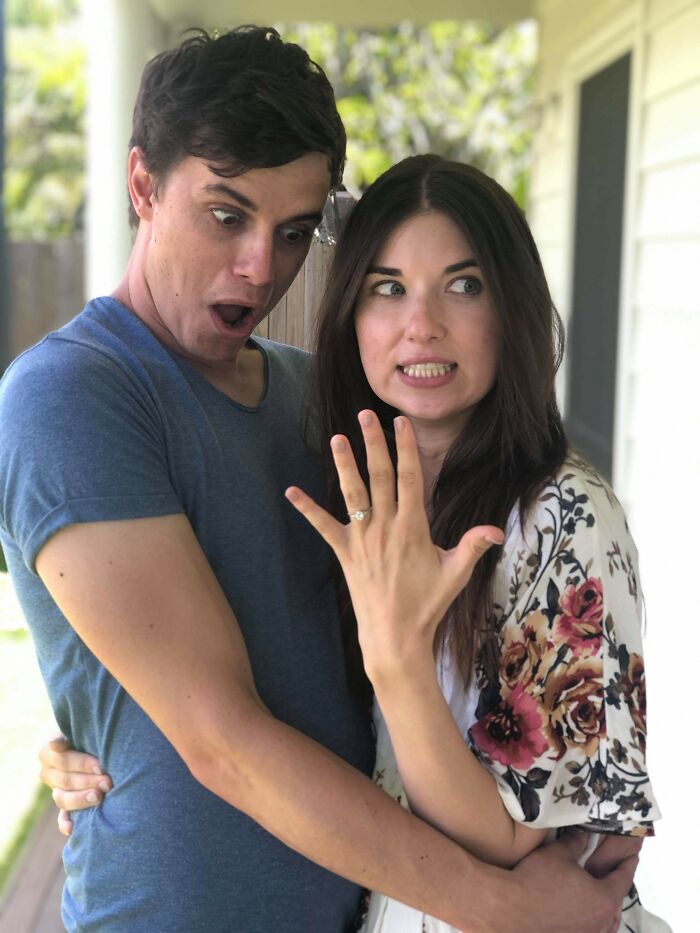 A surprised couple embraces, with the woman displaying an engagement ring, capturing a wholesome moment.