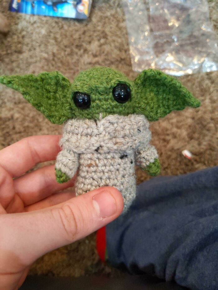 A hand holds a crocheted green creature with large eyes, showcasing a cozy, wholesome Christmas post ambiance.