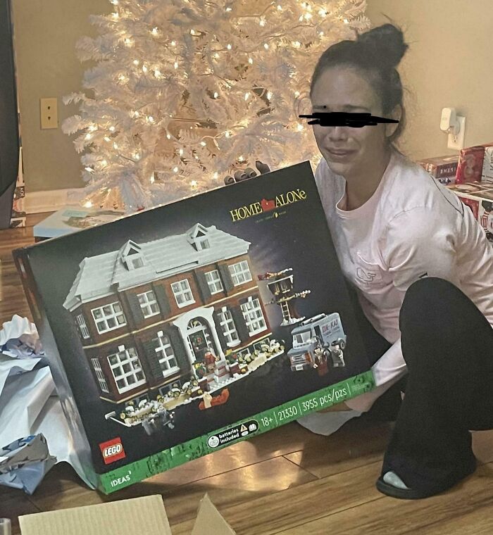 Person joyfully holding a Home Alone LEGO set in front of a lit Christmas tree. Wholesome Christmas setting.