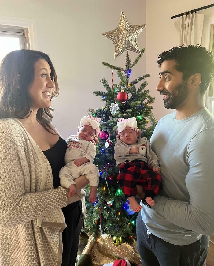 Smiling couple with newborn twins in front of a decorated Christmas tree, capturing a wholesome holiday moment.