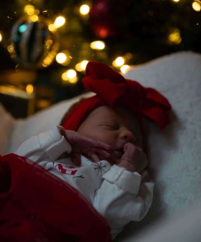 Newborn in festive attire sleeps peacefully near glowing Christmas tree, capturing a wholesome Christmas moment.