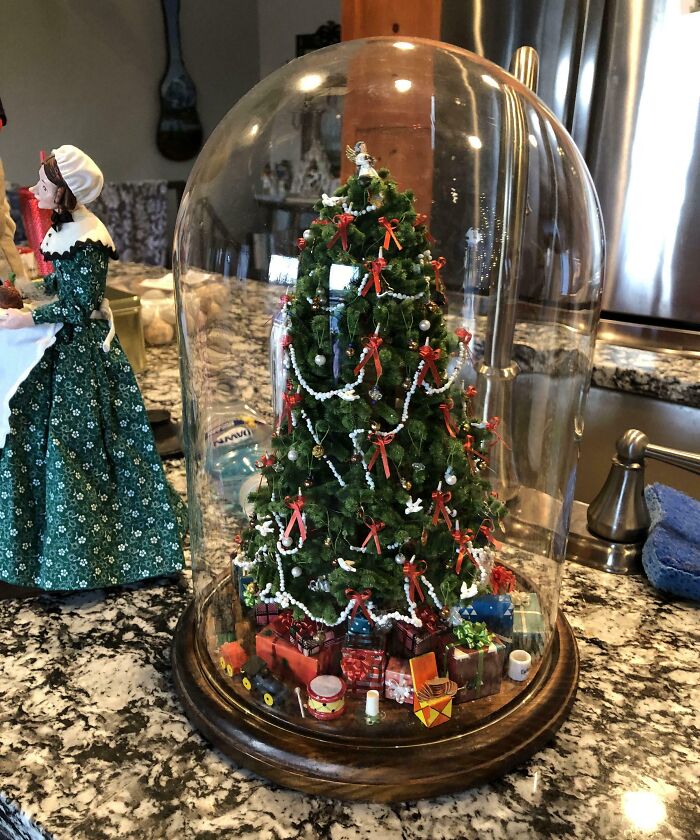 Miniature Christmas tree in a glass dome on a kitchen counter, adorned with red bows and surrounded by tiny gifts.