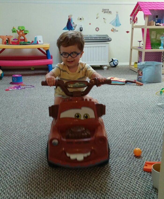 Toddler riding a toy truck indoors, surrounded by Christmas-themed toys and decorations.