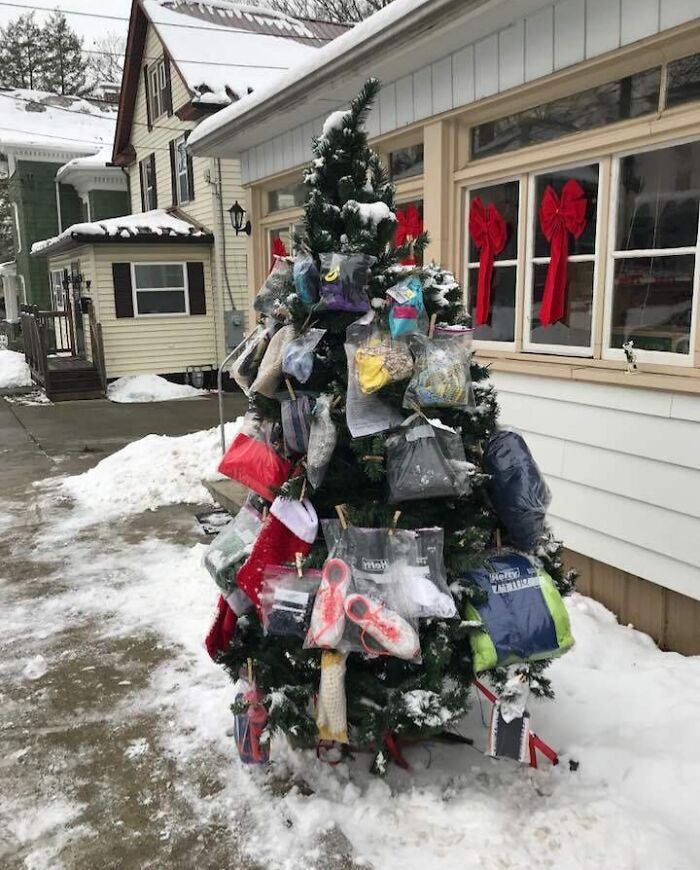 Christmas tree decorated with donated winter essentials, standing outside a snow-covered house with red bows on the windows.