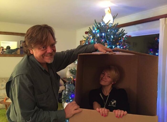 Man smiling next to woman inside a box, with a decorated Christmas tree in the background. Wholesome Christmas setting.