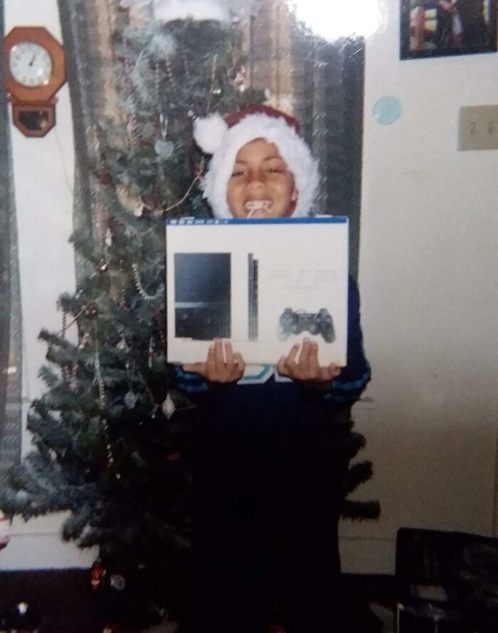 Child in Santa hat joyfully holding a PS2 console box by a Christmas tree, capturing a wholesome Christmas moment.