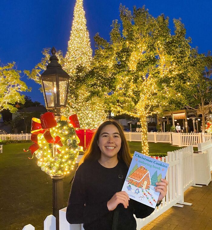 Smiling woman holds a Christmas coloring book in a festive setting with lights and a decorated tree.