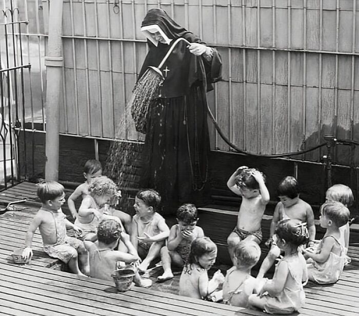 A nun playfully sprays water over a group of laughing children, capturing a lively historical moment.