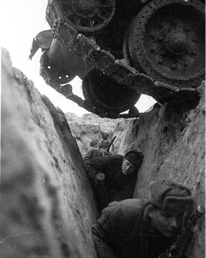 Soldiers in a trench during wartime, with a tank overhead, capturing a historical moment.