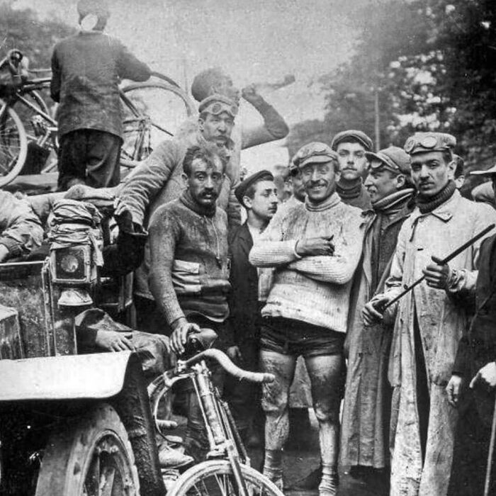 A group of cyclists and crew in vintage attire stand by an old car, showcasing historical moments for a dedicated IG page.