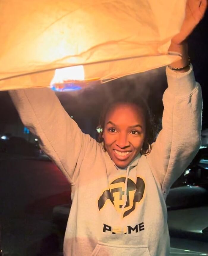 Person holding a lit lantern outdoors, smiling warmly, in a casual setting.