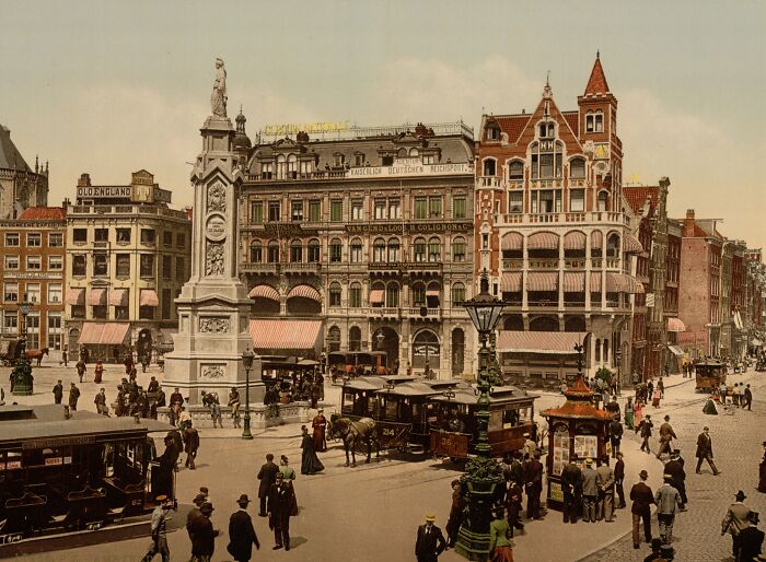 Old color photo of a busy city square from 100 years ago, featuring people, trams, and historical buildings.