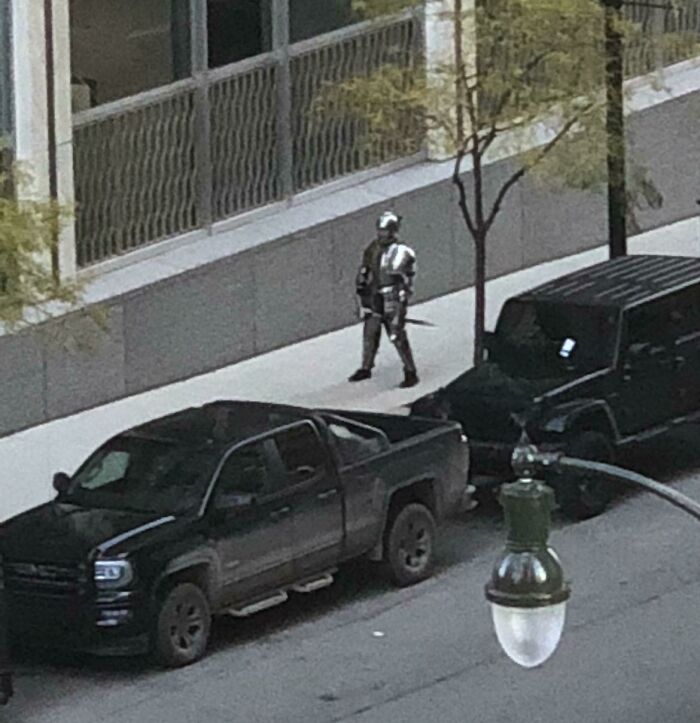 Person in knight armor walks on city street, resembling a scene from a videogame.