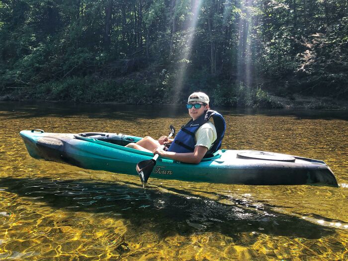 Person paddling a blue kayak on clear water, appearing to float above the surface, as sunlight shines through the trees.