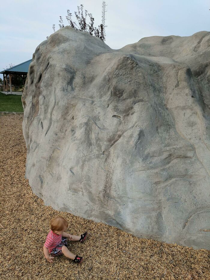 Child sitting near large gray rock that resembles a bad render, surrounded by wood chips in a park.