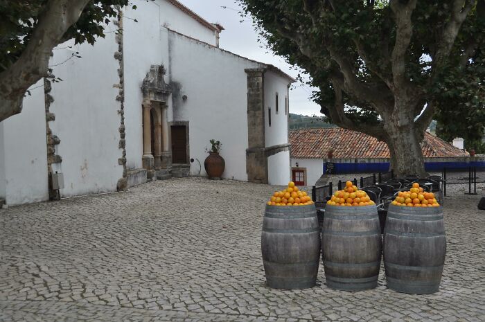 Cobblestone courtyard with barrels of oranges, resembling a bad render with smooth textures and odd proportions.