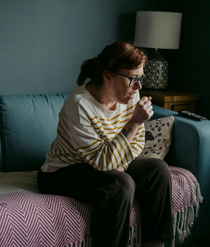 Woman sitting on couch, coughing, wearing glasses and striped top.