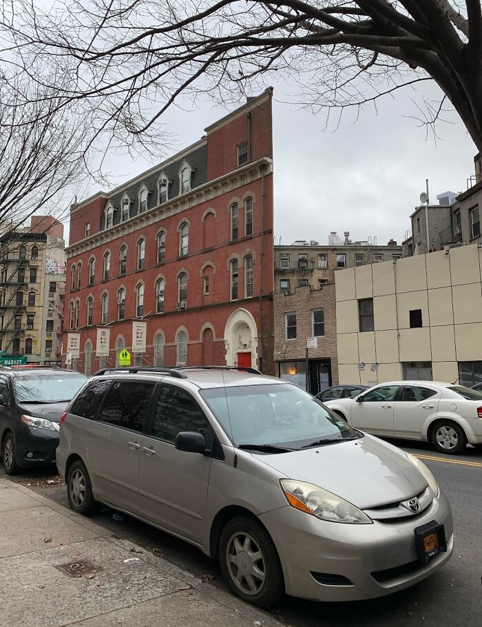 Street scene with a red brick building, tree branches above, and cars parked, resembling a bad 3D render.