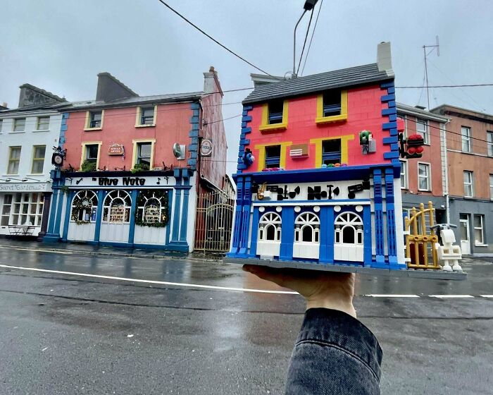 Hand holding a mini LEGO building replica in front of a real building, showcasing cool LEGO builds.