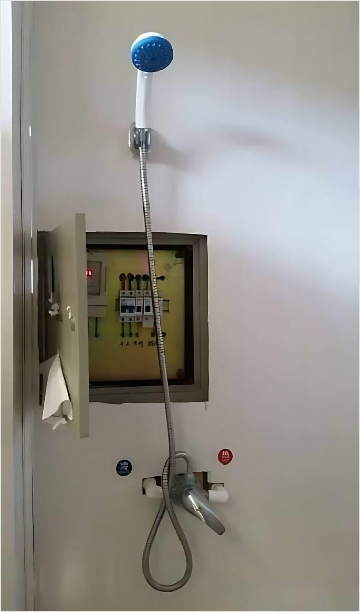 Construction fail showing a showerhead over an exposed electrical panel on a white wall.