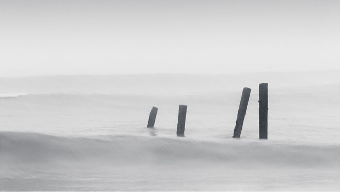 Monochrome photograph capturing waves with wooden posts emerging from the water.