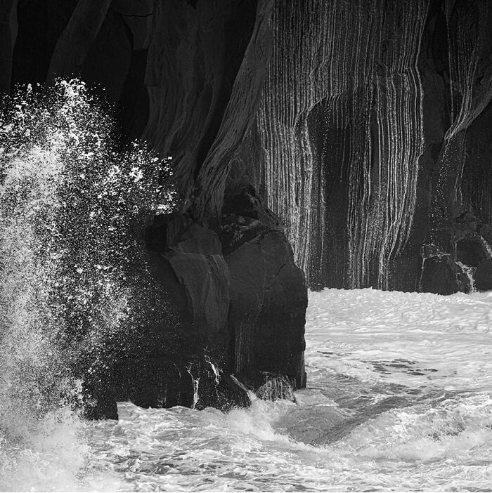 Black and white photo captures waves splashing against rocky cliffs, showcasing fascinating wave photography.