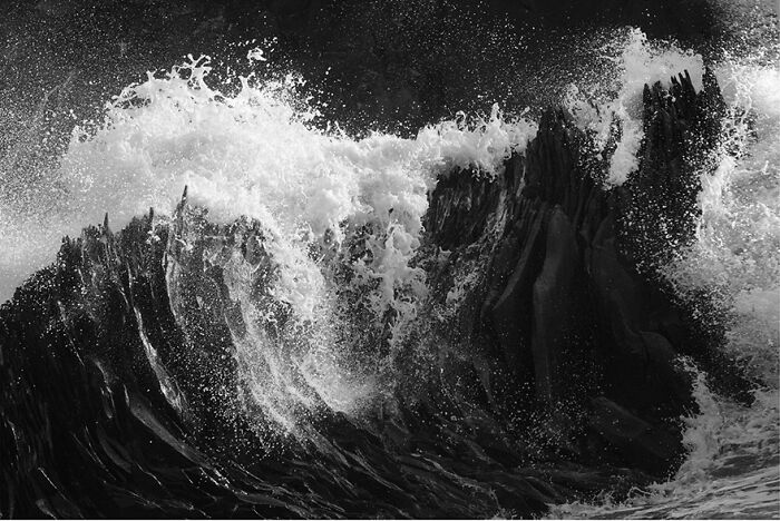 Black and white photo capturing waves crashing dramatically against sharp rocks.