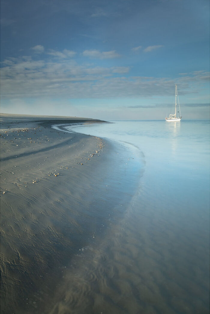 Sailboat on calm water with gentle waves beside a beach at sunset.