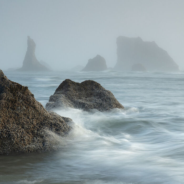 Waves crashing against rocks, captured in a misty, serene seascape by a photographer.