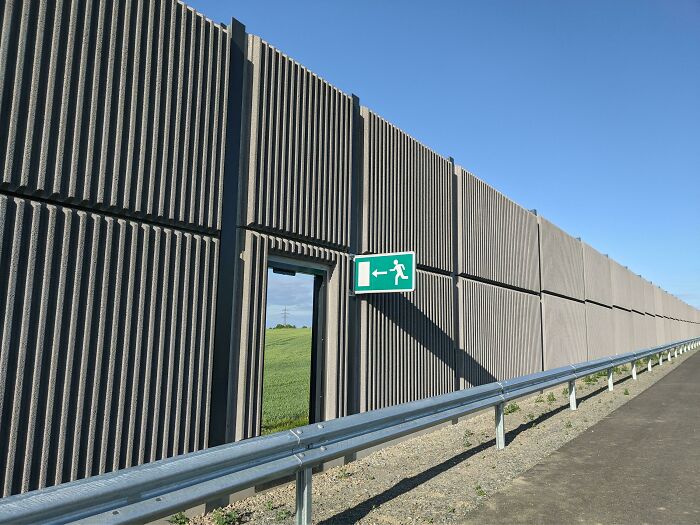 Concrete wall with emergency exit sign, resembling a videogame scene, against a clear blue sky.