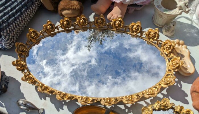 Ornate mirror reflects sky, resembling video game graphics, surrounded by various objects.