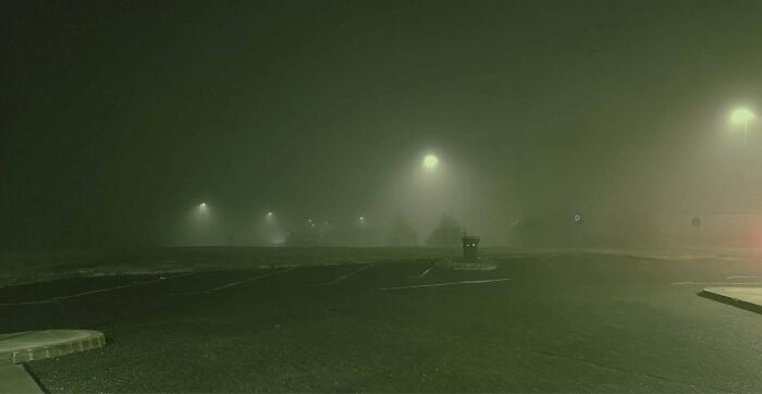 Foggy night scene with streetlights illuminating an empty parking lot, resembling videogame graphics.