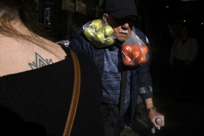 Elderly man in a street market in Israel carrying bags of apples and oranges on his shoulders.