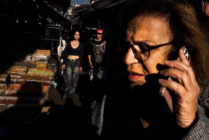 Woman on phone in Israeli street market, with shadowed figures and spice stalls. Israel street photography.