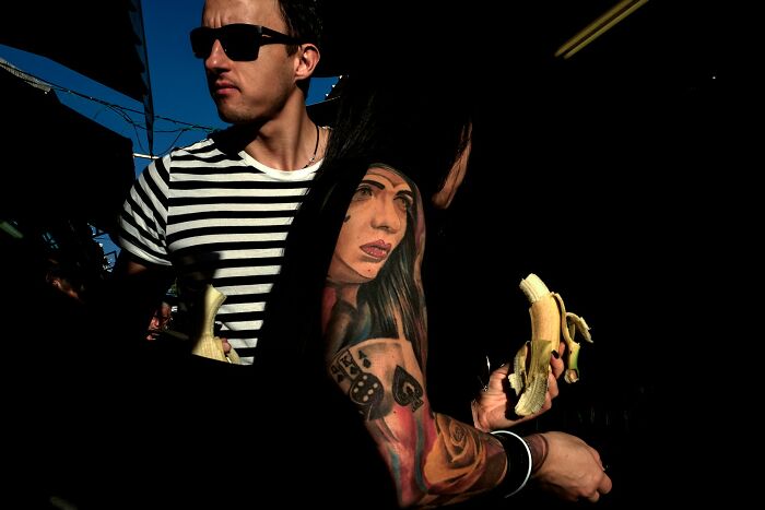 Street scene in Israel: two individuals eating bananas, one with a prominent tattoo on their arm.