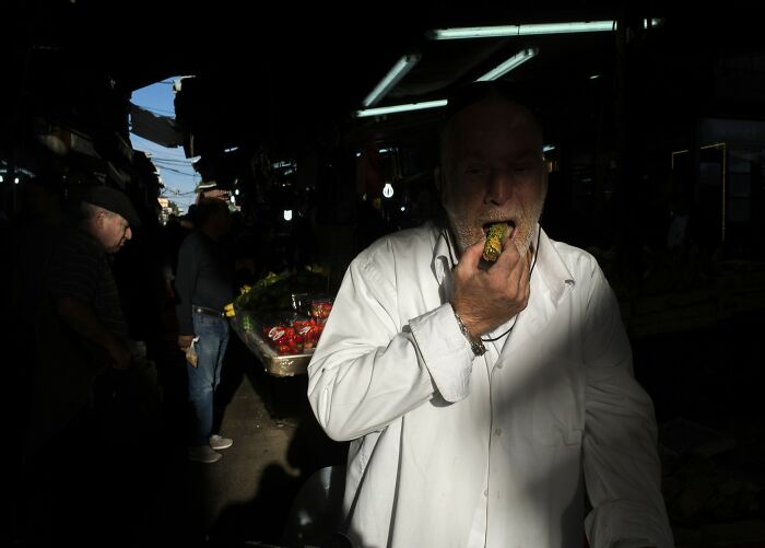 Man in a market eating a cucumber, illuminated by a ray of light, capturing an unedited moment in Israel street life.