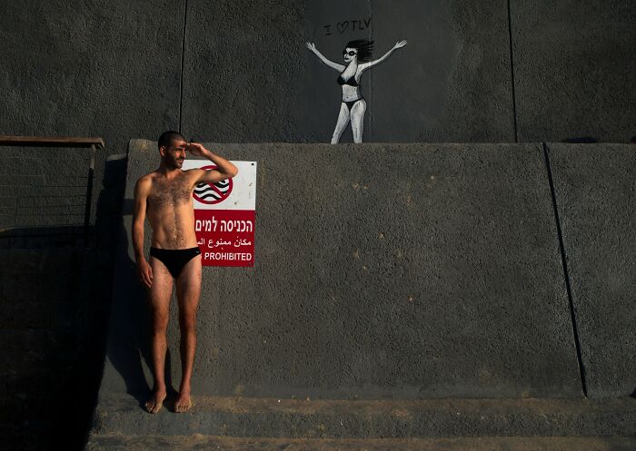 Man in swim trunks stands by a "Swimming Prohibited" sign on a Tel Aviv street, with wall art in the background.